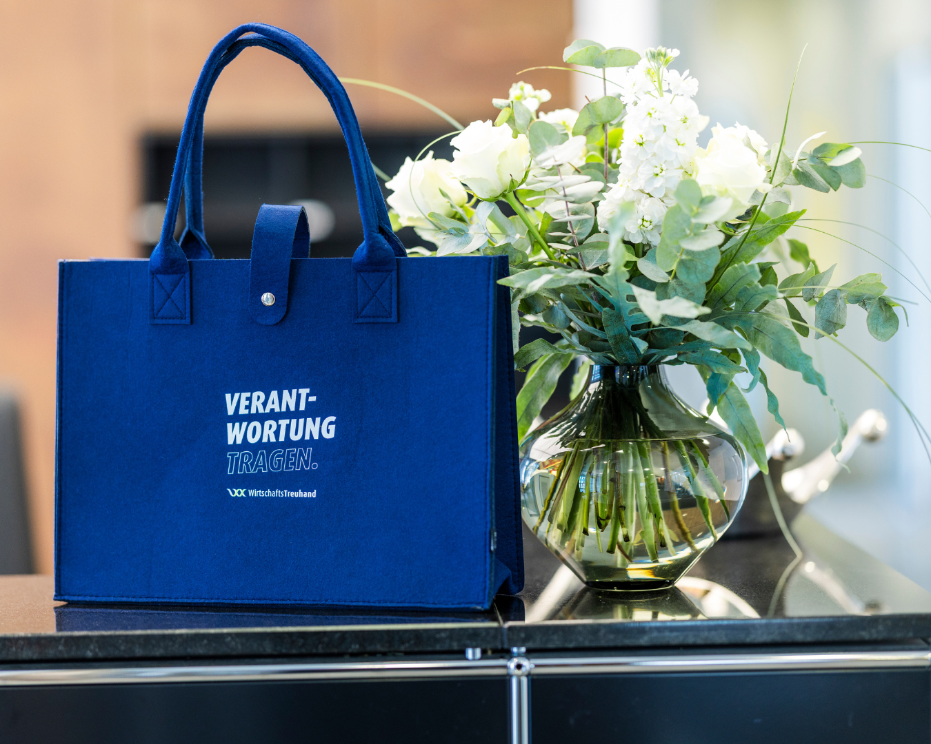 Zu sehen ist eine blaue Tasche auf der steht: "Verantwortung tragen" neben einem weißen Rosenstraß in einer Vase.